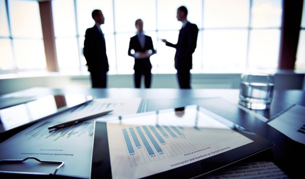 Close-shot of a tablet computer displaying financial data, three businessmen standing in the background