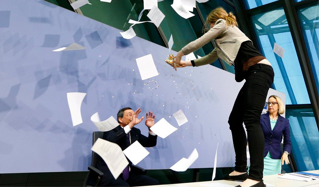 Uma mulher em protesto contra o Banco Central Europeu subiu em uma mesa na frente do presidente do BCE, Mario Draghi, durante entrevista coletiva nesta quarta-feira; "Fim da ditadura do BCE", gritava a mulher sobre a mesa, enquanto jogava confetes e pedaços de papel em cima de Draghi