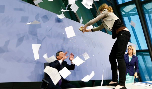 Uma mulher em protesto contra o Banco Central Europeu subiu em uma mesa na frente do presidente do BCE, Mario Draghi, durante entrevista coletiva nesta quarta-feira; "Fim da ditadura do BCE", gritava a mulher sobre a mesa, enquanto jogava confetes e pedaços de papel em cima de Draghi