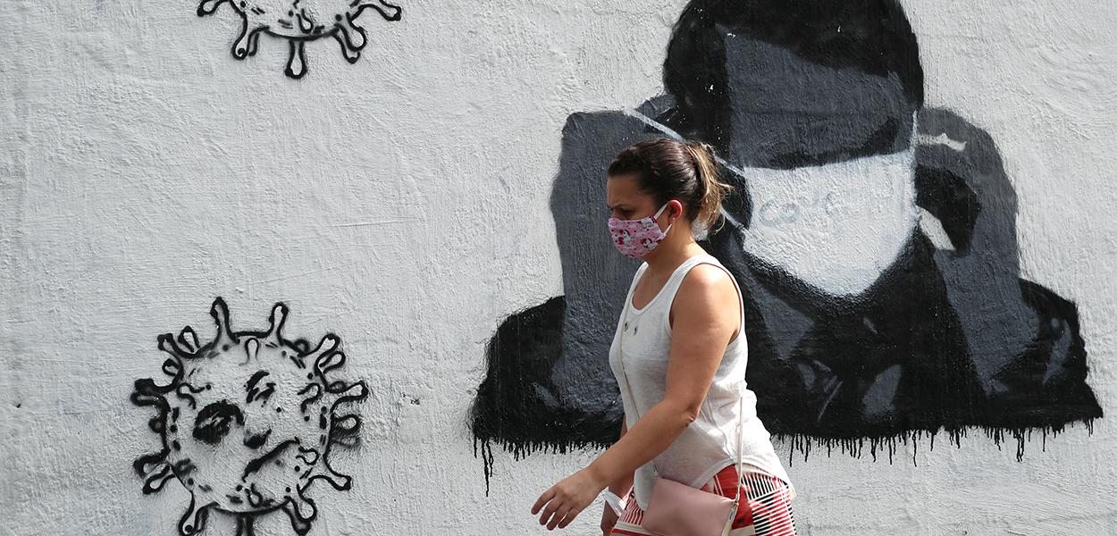 Mulher passa por grafite do presidente Jair Bolsonaro ajustando sua máscara protetora e em forma de vírus, em meio ao surto de Covid-19 no Rio de Janeiro.