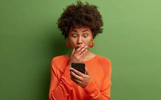 Horizontal shot of amazed adult woman being deeply surprised, stares at smartphone display, reads shocking news on website, dressed in orange jumper, has bugged eyes, covers mouth. Omg, its horrible!