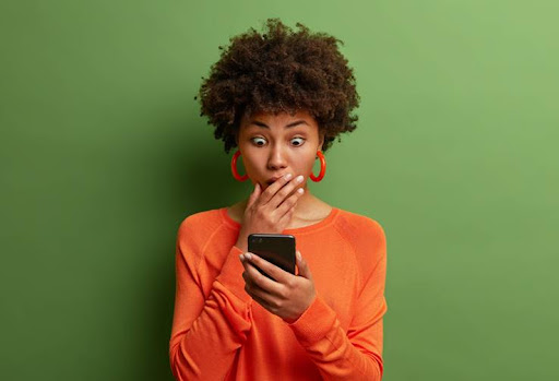 Horizontal shot of amazed adult woman being deeply surprised, stares at smartphone display, reads shocking news on website, dressed in orange jumper, has bugged eyes, covers mouth. Omg, its horrible!