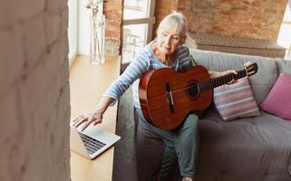 Learning to play guitar online. Senior woman studying at home, getting online courses, self-development. Caucasian woman using modern devices for education, spending time for new job or hobby.