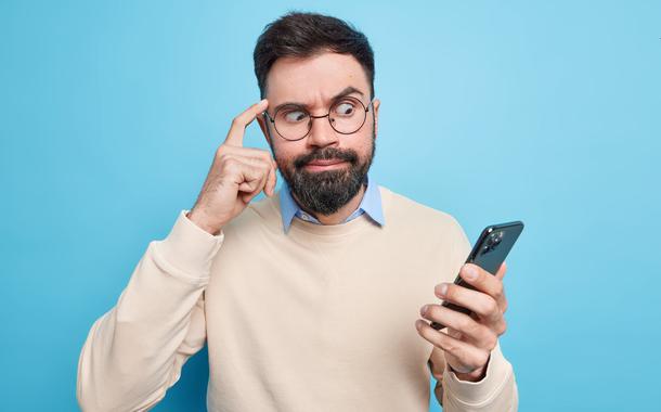 Embarrassed bearded man focused at smartphone keeps finger on temple tries to concentrate on information stares at display dressed in neat sweater poses against blue background. Technology concept