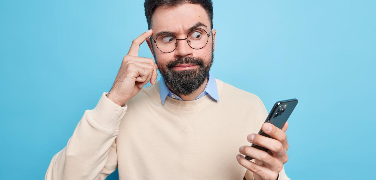 Embarrassed bearded man focused at smartphone keeps finger on temple tries to concentrate on information stares at display dressed in neat sweater poses against blue background. Technology concept