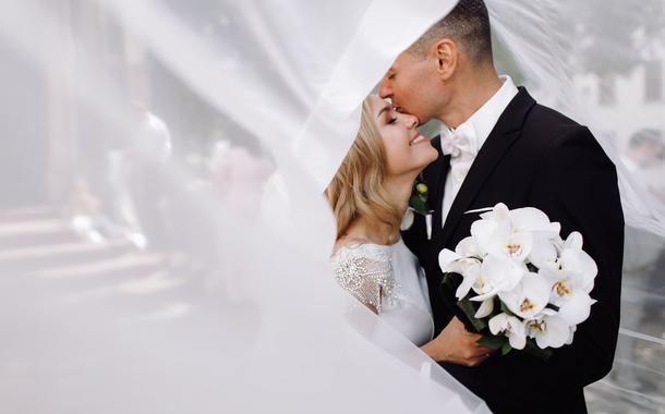 Groom in black tuxedo hugs tender stunning bride while they stand on the street of old European town