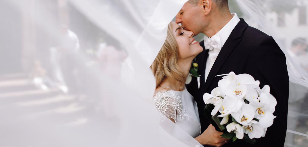 Groom in black tuxedo hugs tender stunning bride while they stand on the street of old European town