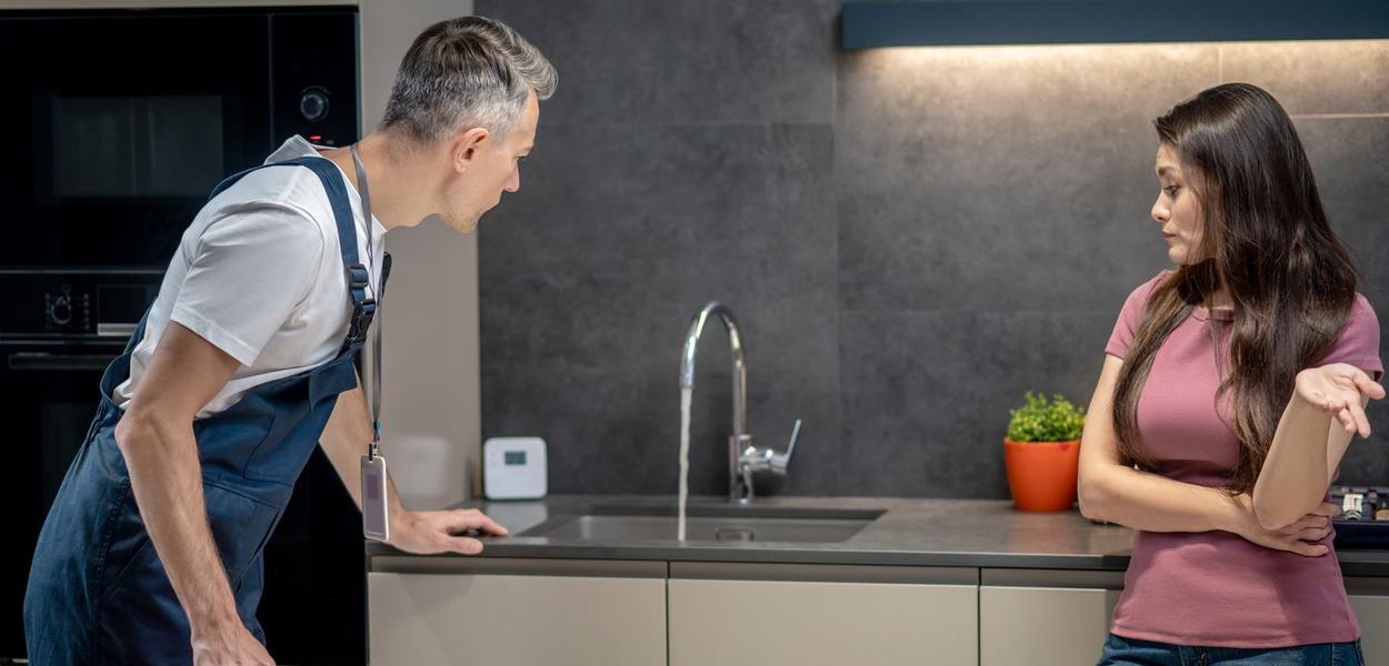 Breakdown, plumbing. Pretty dark haired middle aged woman in tshirt with question gesture and male plumber in overalls with special case looking at broken faucet in kitchen