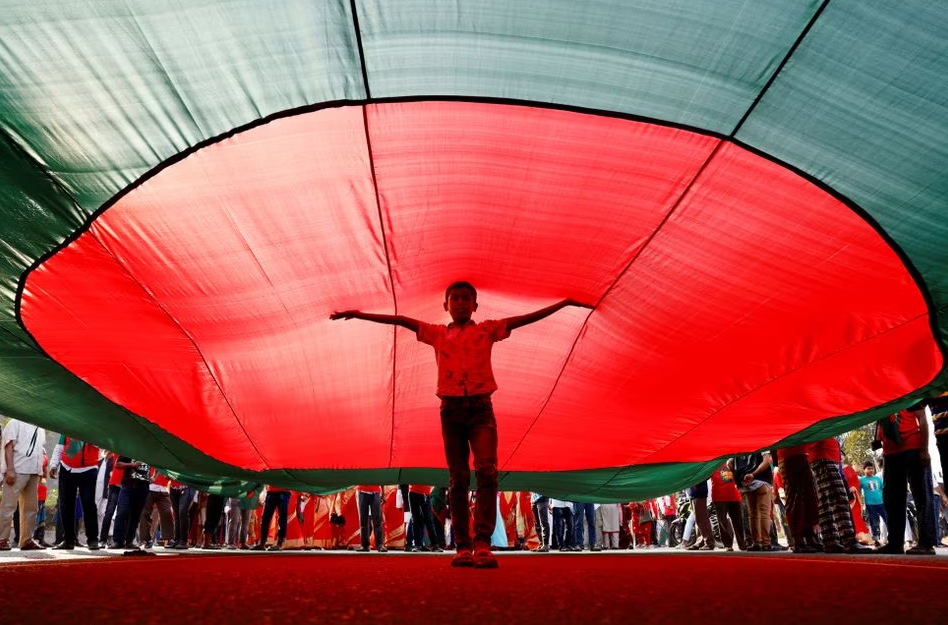 Um menino posa para uma foto sob uma enorme bandeira de Bangladesh, durante um comício três dias antes do 50º aniversário do Dia da Vitória do país, em frente ao prédio do parlamento em Dhaka, Bangladesh, em 13 de dezembro de 2021