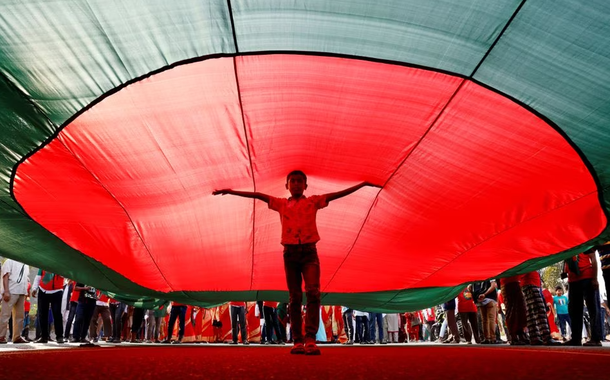 Um menino posa para uma foto sob uma enorme bandeira de Bangladesh, durante um comício três dias antes do 50º aniversário do Dia da Vitória do país, em frente ao prédio do parlamento em Dhaka, Bangladesh, em 13 de dezembro de 2021